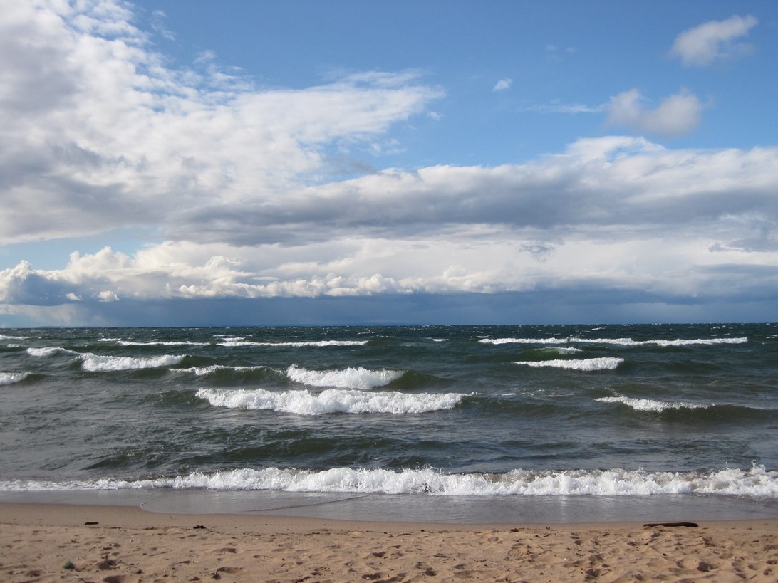 En naturskjønn utsikt over havet med bølger som slår inn mot en sandstrand under en dramatisk himmel.