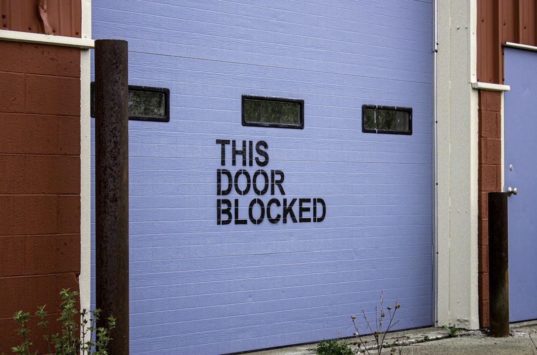 En blå garasjeport med teksten 'THIS DOOR BLOCKED' malt i sort.